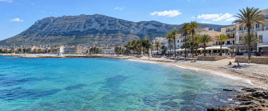 Clear turquoise water over dark rocks in a curving bay, palm trees and white buildings along the shore and a long grey limestone mountain behind